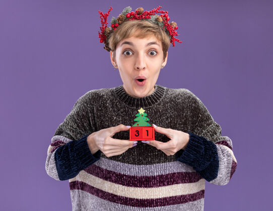 头令人印象深刻的年轻漂亮女孩戴着圣诞花环手持圣诞树玩具与日期隔离在紫色墙上年轻穿着日期