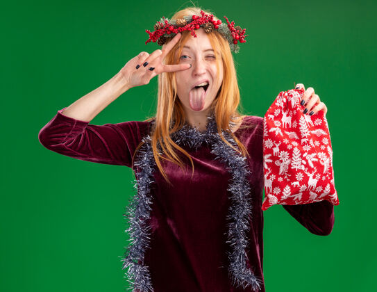 眨眼快乐的眨着眼睛的年轻漂亮的女孩穿着红色的衣服 脖子上戴着花环和花环 拿着圣诞袋 露出舌头 做着与世隔绝的绿色墙壁上的和平手势花环花环和平