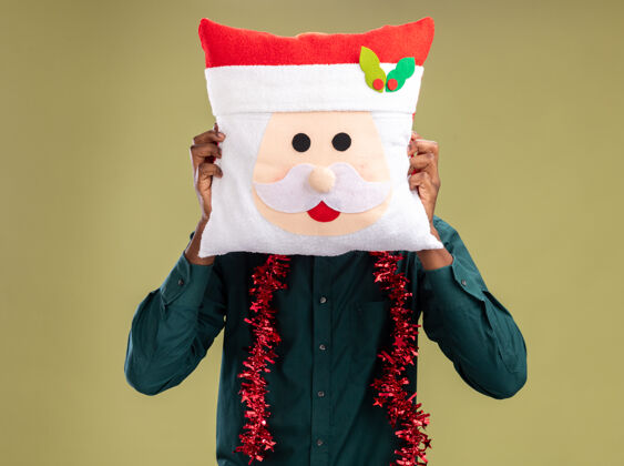 非洲戴着花环的非洲裔美国人拿着圣诞枕头 把脸藏在枕头后面 站在绿色的背景上站枕头脸