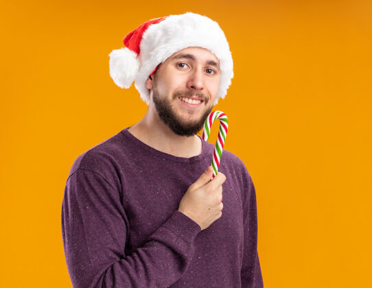 毛衣一个穿着紫色毛衣 戴着圣诞帽 手持糖果手杖的快乐年轻人站在橙色背景下 面带微笑地看着相机站着圣诞老人帽子