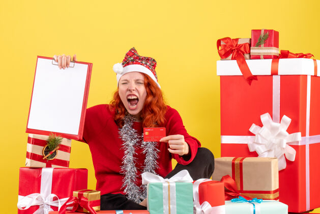 盒子前视图年轻女子坐在圣诞礼物周围拿着银行卡在黄色的墙上礼物情感帽子