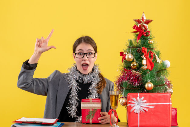 节日前视图：戴眼镜的年轻女孩坐在桌子旁 手指指向圣诞树和鸡尾酒礼物礼物年轻女孩桌子