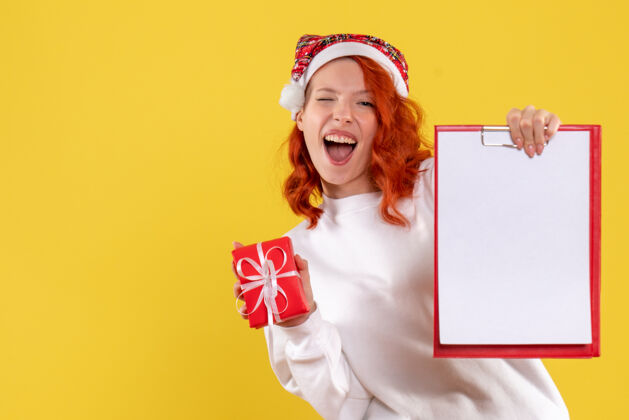 年轻手持小圣诞礼物和黄色墙上的文件笔记的年轻女子的正面视图小便条礼物