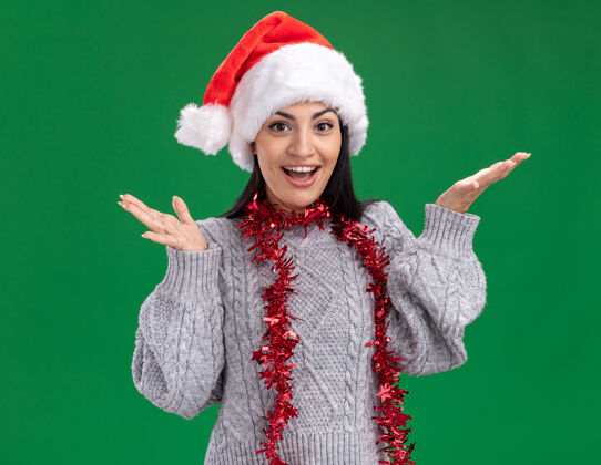 空年轻的白人女孩戴着圣诞帽 脖子上戴着金箔花环 空荡荡的双手孤立在绿色的墙上脖子年轻花环