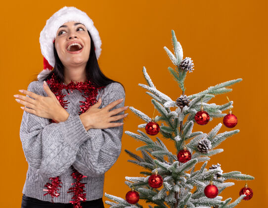 圣诞节令人印象深刻的年轻白人女孩戴着圣诞帽 脖子上戴着金箔花环 站在装饰好的圣诞树旁 双手交叉放在胳膊上 孤立地看着橙色的墙上金属丝帽子保持