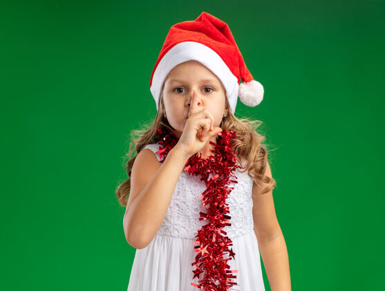 圣诞节看起来小女孩戴着圣诞帽 脖子上戴着花环 在绿色的墙上表现出沉默的姿态穿着脖子花环