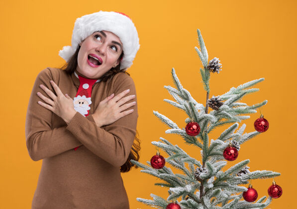戴着快乐的仰望着戴着圣诞帽打着领带的年轻漂亮女孩站在圣诞树旁把手放在肩膀上孤立地站在橙色的墙上年轻帽子放着