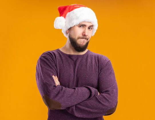 表情穿着紫色毛衣 戴着圣诞帽的年轻人抱着怀疑的表情看着摄像机 双臂交叉着站在橙色的背景上交叉圣诞老人年轻