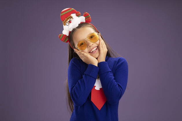 边缘小女孩穿着蓝色高领毛衣 系着红色领带 头上戴着有趣的圣诞戒指 站在紫色的背景下 看着镜头 脸上洋溢着幸福的笑容领带头小