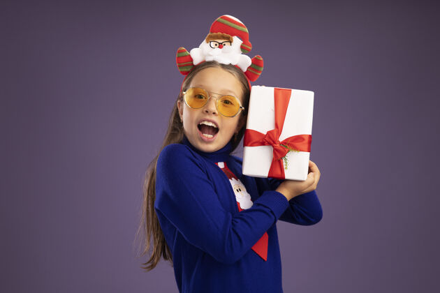 领带小女孩穿着蓝色高领毛衣 打着红色领带 头上戴着有趣的圣诞戒指 手里拿着礼物 站在紫色的背景下 开心而兴奋地看着相机女孩礼物搞笑
