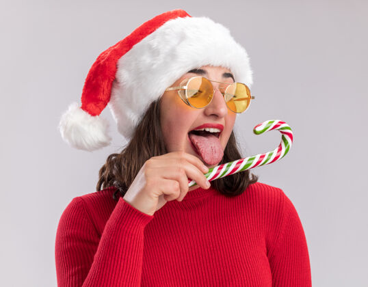 站着穿着红色毛衣 戴着圣诞帽 戴着眼镜 拿着糖果手杖的年轻女孩站在白色的背景下 试图品尝它的快乐和喜悦女孩毛衣快乐