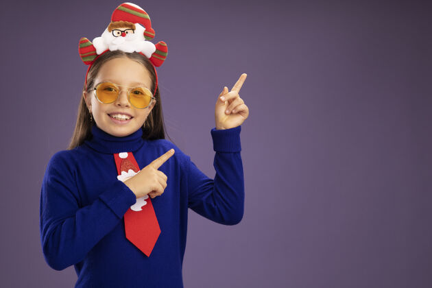 站着小女孩穿着蓝色高领毛衣 打着红色领带 头上戴着有趣的圣诞戒指 看着摄像机 兴高采烈地笑着 食指指向站在紫色背景上的一边指点微笑女孩