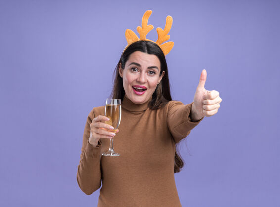 头发微笑着的年轻漂亮女孩 穿着棕色毛衣 戴着圣诞发箍 手里拿着一杯香槟 在蓝色的背景上孤立地竖起大拇指玻璃毛衣展示