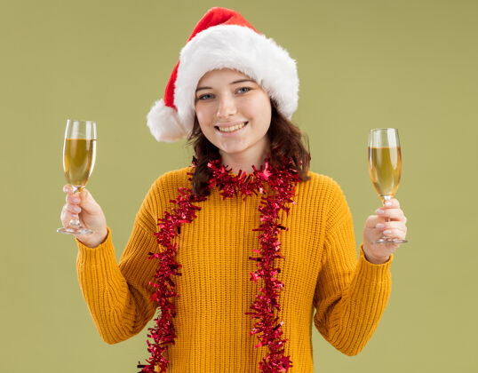 橄榄微笑的斯拉夫女孩戴着圣诞帽 脖子上戴着花环 手里拿着一杯香槟酒 背景是橄榄绿 还有复印空间花环眼镜脖子