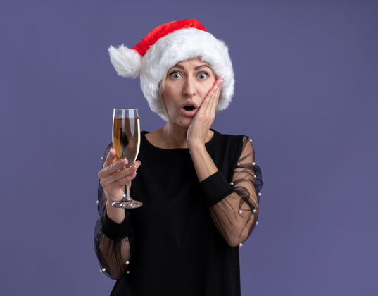 香槟关心的中年金发女人戴着圣诞帽 拿着香槟酒杯 手放在脸上 隔离在紫色的墙上 留着复制空间关心金发穿着