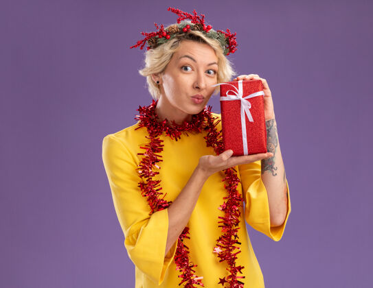 脖子令人印象深刻的年轻金发女子头戴圣诞花环和金箔花环在脖子上拿着礼品包看着噘起的嘴唇隔离在紫色的墙壁与复制空间金发金箔花环