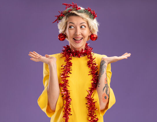 女人令人印象深刻的年轻金发女子头戴圣诞花环 脖子上戴着金箔花环 一边看一边空空的手 耳朵上挂着圣诞饰品 孤立地挂在紫色的墙上空侧面花环