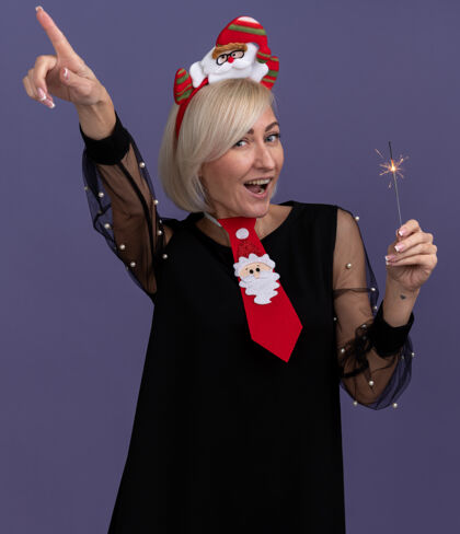 中年令人印象深刻的中年金发女人戴着圣诞老人的头带和领带举行节日火花指向孤立的紫色墙上圣诞老人圣诞节领带