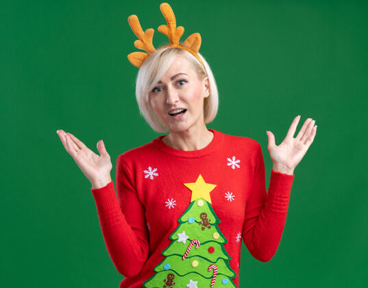 穿着令人印象深刻的中年金发女人穿着圣诞驯鹿鹿角头带和圣诞毛衣显示空手孤立在绿色的墙上女人印象深刻毛衣