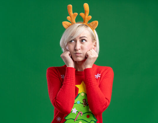 保持无聊的中年金发女人戴着圣诞驯鹿鹿角头带和圣诞毛衣看着一边鼓鼓的脸颊保持手放在脸上隔离在绿色的墙上复制空间女人金发穿