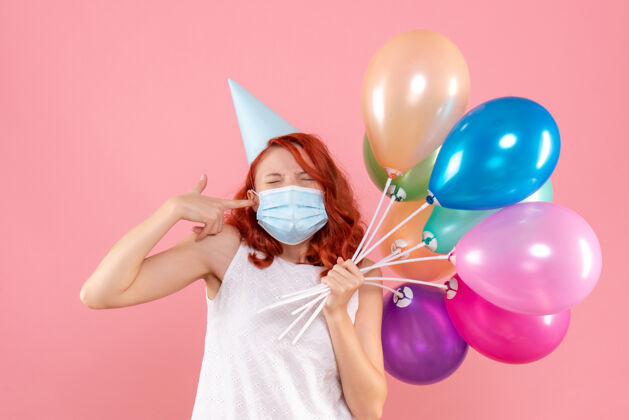 前面前视图中的年轻女子拿着五颜六色的气球在无菌面具上粉红色的墙壁举行病毒气球