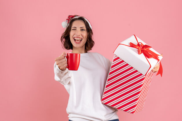 人年轻女子手持圣诞礼物和粉红色墙上的一杯茶的前视图女人圣诞节女性