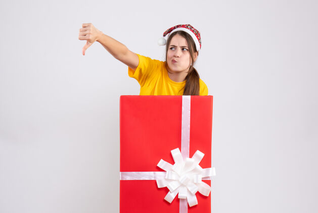 大戴着圣诞帽的快乐女孩站在白色大圣诞礼物后面做拇指朝下的标志女孩制造圣诞节