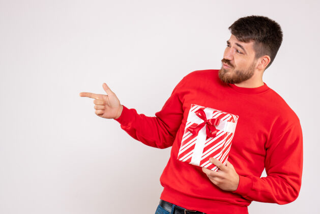 男性身穿红衫的年轻人手持圣诞礼物站在白墙上的正视图现在成人圣诞节