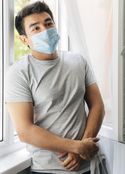 社会大流行期间 家里戴着医用口罩的男人坐在窗户旁边流行病隔离预防