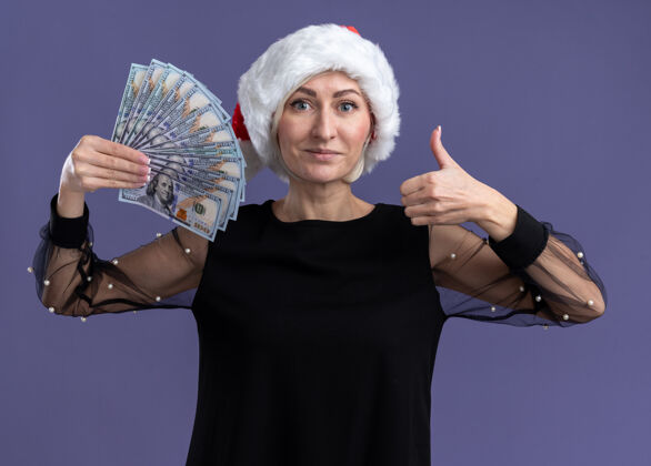 金发高兴的中年金发女人戴着圣诞帽拿着钱看着相机显示拇指向上孤立的紫色背景帽子看高兴
