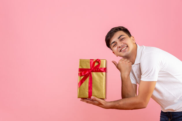 微笑一个年轻人在粉红色的墙上给别人送圣诞礼物的正面图盒子成人十二月