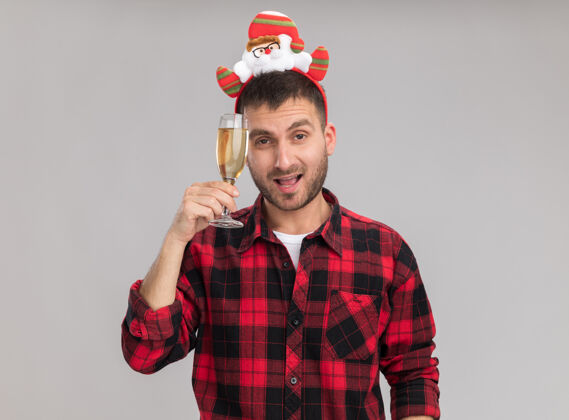 头自信的白人年轻人戴着圣诞头带 拿着一杯香槟 用它抚摸着头 看着隔离在白色背景上的相机看着年轻圣诞