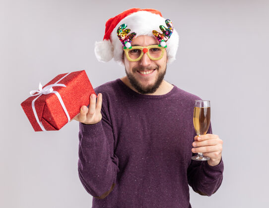 年轻快乐的年轻人穿着紫色毛衣 戴着圣诞帽 戴着滑稽的眼镜 手里拿着一杯香槟和礼物眼镜圣诞快乐圣诞老人