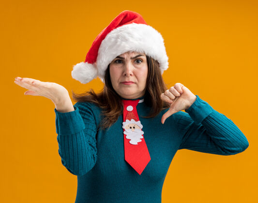 帽子未租成年白人妇女与圣诞帽和圣诞领带拇指朝下 保持开放的手在橙色背景与复制空间隔离领带手橙色