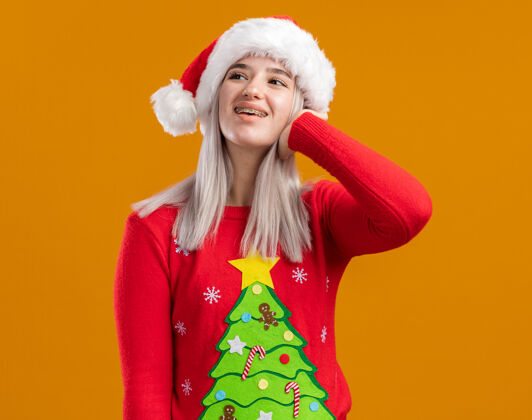 金发穿着圣诞毛衣 戴着圣诞帽的金发女郎站在橙色背景下 开心地看着一旁毛衣圣诞老人帽子