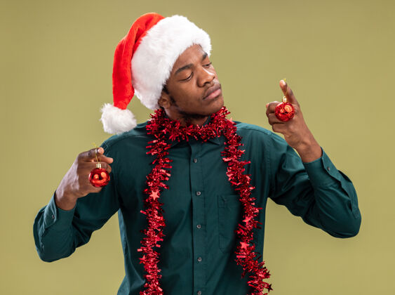 非洲戴着圣诞帽 戴着花环 拿着圣诞球的非裔美国人站在绿色背景下严肃地看着球站着圣诞节脸