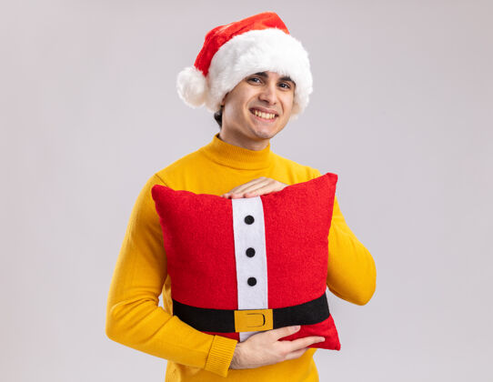高领毛衣快乐的年轻人穿着黄色高领毛衣 戴着圣诞帽 抱着圣诞枕头 站在白色背景下 面带微笑地看着相机圣诞老人脸枕头