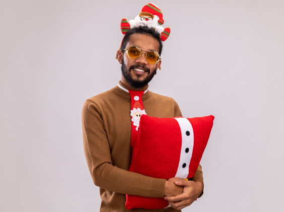 轮辋快乐的非洲裔美国人 身穿棕色毛衣 头戴圣诞老人戒指 打着有趣的红色领带 抱着圣诞枕头 站在白色背景下 面带微笑地看着相机脸美国人枕头
