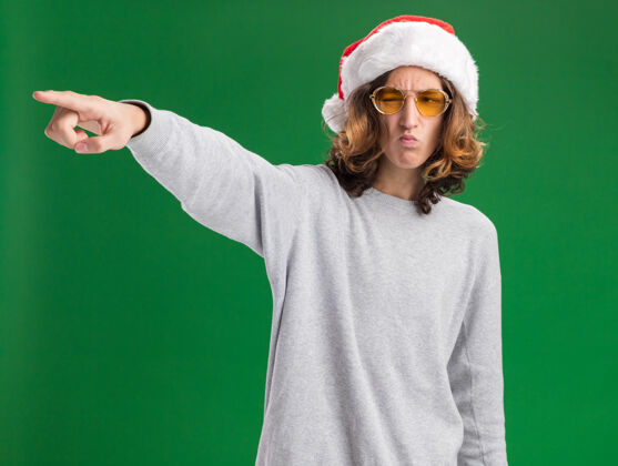 眼镜戴着圣诞老人帽和黄色眼镜的年轻人 眉头紧锁 用食指指着站在绿色背景上的一边指着圣诞节食指