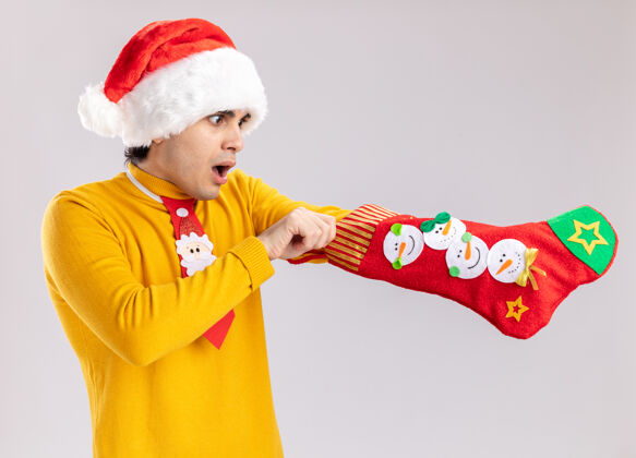 搞笑一个穿着黄色高领毛衣 戴着圣诞老人帽 打着滑稽领带 手里拿着圣诞长袜的年轻人站在白色的背景下 看上去既惊讶又惊讶高领毛衣长袜站着