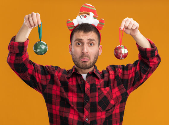 男人无知的白人年轻人戴着圣诞老人的头带 手里拿着圣诞饰品孤立在橙色的墙上戴着不知所措圣诞饰品
