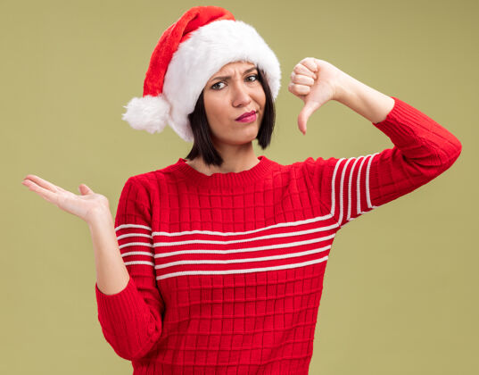未出租戴着圣诞帽的未被释放的年轻女孩看着相机 在橄榄绿的背景下 她双手空空 拇指朝下圣诞老人橄榄空