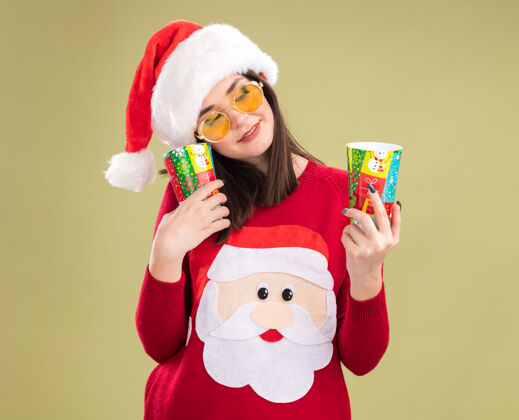 帽子高兴的年轻漂亮的白人女孩穿着圣诞老人毛衣和帽子戴着眼镜拿着塑料圣诞杯看着其中一个孤立的橄榄绿背景圣诞节橄榄年轻