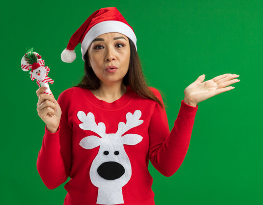 毛衣戴着圣诞老人帽 穿着红色毛衣的年轻女子手持圣诞糖果手杖 困惑地看着摄像机 举起手臂站在绿色背景上手杖困惑年轻人