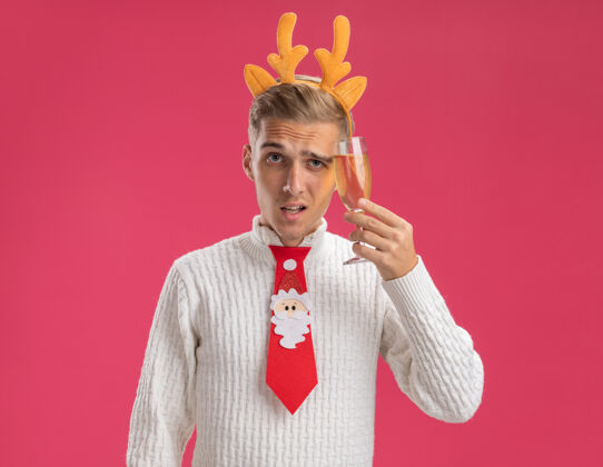 抚摸可疑的年轻帅哥戴着驯鹿的鹿角头带 打着圣诞老人的领带 手里拿着一杯香槟 把它孤立在粉红色的墙上驯鹿圣诞节香槟