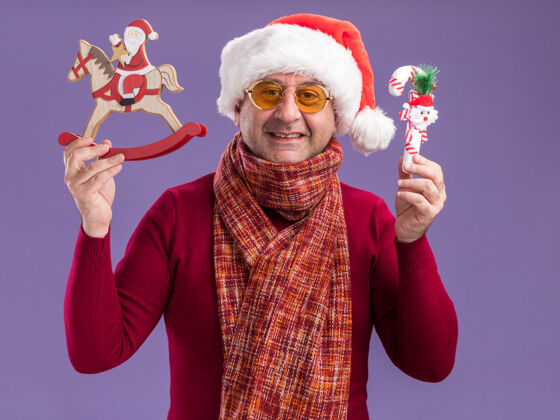 站着中年男子戴着圣诞老人帽 脖子上围着暖和的围巾 戴着黄色眼镜 站在紫色的背景下 展示着圣诞玩具 快乐欢快的微笑微笑玩具脖子