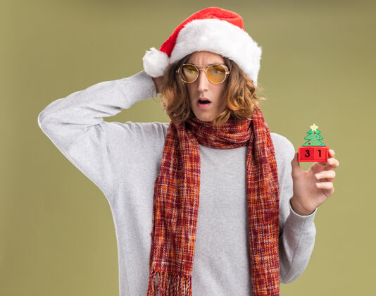 日期戴着圣诞老人帽 戴着黄色眼镜 脖子上围着暖和的围巾的年轻人 手里拿着印有新年日期的玩具立方体 站在绿色的背景下焦急地看着一边站着脖子围巾
