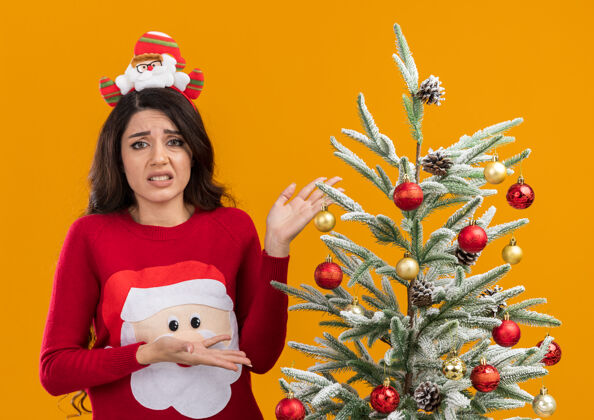 站着恼怒的年轻漂亮女孩戴着圣诞老人的头带和毛衣站在装饰过的圣诞树旁指着它看着橙色背景上孤立的摄像机橙色恼火头带