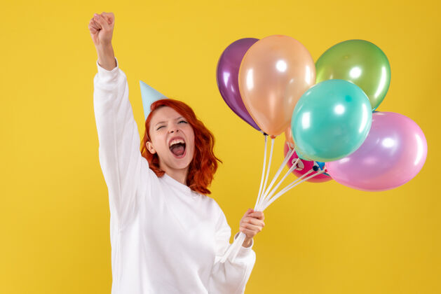 庆祝黄色墙壁上手持可爱彩色气球的年轻女子的正视图气球前面礼物
