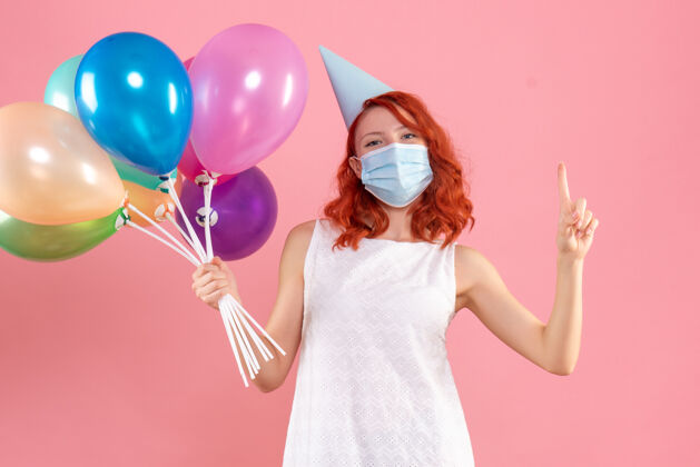快乐前视图中的年轻女子拿着五颜六色的气球在无菌面具上粉红色的墙壁庆祝不育女性